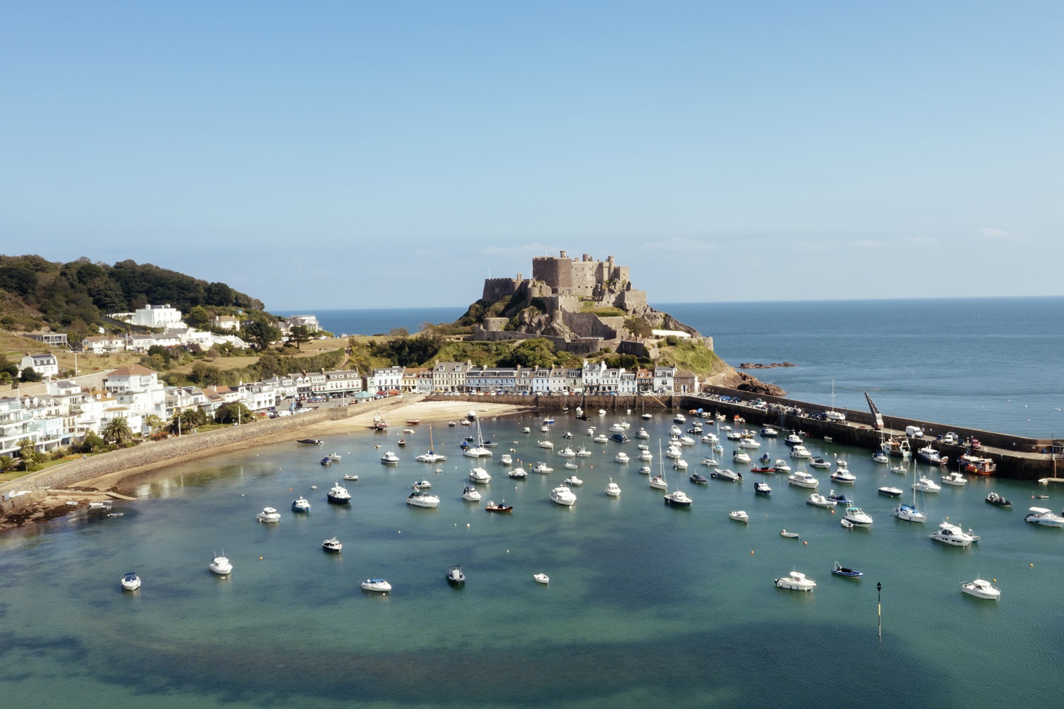Luftaufnahme der Burg Mont Orgueil, die auf einer felsigen Halbinsel über dem Hafen von Gorey mit vielen weißen Booten auf Jersey thront. Ein Dorf säumt die Bucht.