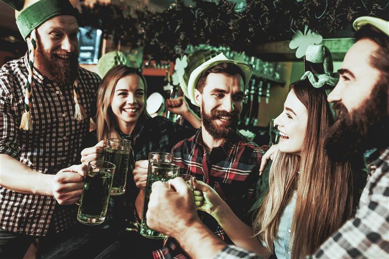 Fünf lachende junge Erwachsene feiern in einem Pub, geschmückt mit grünen Kleeblättern, und stoßen mit grünen Bieren an. Einige tragen St. Patrick's Day Hüte.